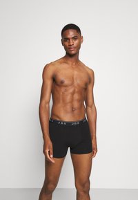 Svarta boxershorts med en märkestextil i midjan, med ett slätt, stretchigt tyg och en figurnära design, modellen står upprätt utan tröja.