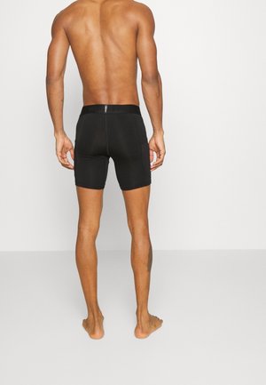 Ein Mann steht barfuß und mit dem Rücken zur Kamera, trägt schwarze Shorts bis zur Mitte des Oberschenkels mit seitlichen Taschen vor einem schlichten Hintergrund.