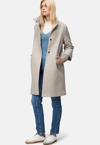 Manteau en laine beige avec grand col et fermetures à boutons, associé à un gilet en tricot bleu et un jean slim. Des baskets blanches complètent la tenue.