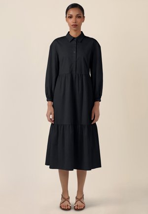 Woman standing wearing a long-sleeve black tiered midi dress with buttons and ankle-strap sandals, neutral beige background.