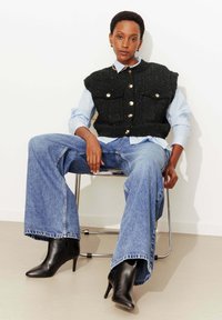 Veste noire texturée avec des boutons dorés, chemise bleu clair en dessous, associée à un jean bleu à jambes larges et des bottines noires pointues. Assise sur une chaise.