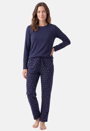 Donna in piedi a piedi nudi che indossa una maglietta blu scuro a maniche lunghe e pantaloni del pigiama blu scuro con un piccolo motivo a cuori bianchi su sfondo uniforme.