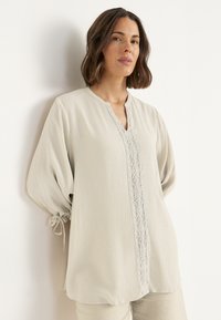 Vrouw met schouderlang donker haar, gekleed in een beige gebreide blouse met kantdetails en strikmouwen, staande tegen een witte muur.