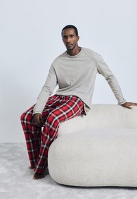 Pelēka garām piedurknēm krekls, sarkanā un melnā pleda pidžamas bikses, sēžot uz mīkstas baltas izliekta dīvāna, kailas kājas uz gaiša paklāja.