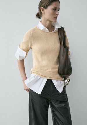 Donna che indossa un maglione beige a maglia sopra una camicia bianca e pantaloni neri, con una borsa a tracolla in pelle marrone scuro sulla spalla.