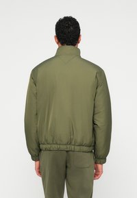 Άνδρας με την πλάτη γυρισμένη, φορώντας καπαρντίνα ελαιόχρωμου και αντίστοιχα παντελόνια, σε απλό λευκό φόντο.