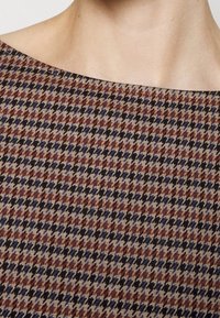 Gros plan d'une personne portant un vêtement à col montant avec un petit motif pied-de-poule dans des tons bleus, marron et beige.