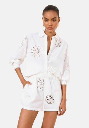 Woman wearing white button-up shirt and shorts set with sun and spiral cutout patterns, standing with one hand in pocket against plain background.