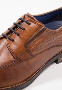 Chaussure en cuir marron à finition lisse, avec des lacets noirs et des accents perforés. Elle présente un intérieur bleu contrastant et une semelle durable.