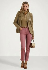 Mulher vestindo um blazer bege e uma polo, calças cor-de-rosa, cinto castanho e sandálias, segurando uma pequena bolsa castanha e bege, de pé contra um fundo liso.