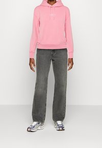 Rosa huvtröja med framsidan logo, tillsammans med grå utsvängda jeans och flerfärgade sneakers. Material i bomull med en slät textur.