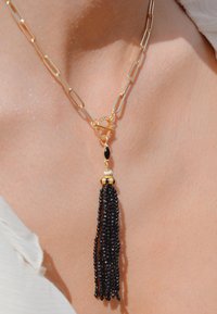 Collier chaîne ton or avec un pendentif ovale et un long gland à perles noires surmonté d'un capuchon décoratif en or.