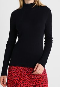 Pull en col roulé noir à côtes avec des manches longues, présentant un design ajusté. Porté par-dessus une jupe rouge à motif léopard.