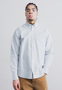 Light blue long-sleeve button-up shirt featuring a chest pocket, straight collar, and subtle texture, paired with brown corduroy trousers.