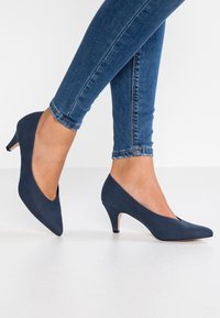Chaussures à talons aiguilles bleu marine avec un bout pointu et un design en forme de V. Texture lisse, talon incurvé et style minimaliste. Convient pour une tenue formelle.