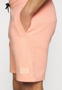 Shorts couleur pêche en tissu doux, avec des poches latérales et une petite étiquette de marque sur l'ourlet. Détail de la ceinture à cordon visible.