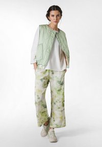 Hellgrüne gesteppte Weste mit rundem Ausschnitt, kombiniert mit weißen, locker sitzenden Hosen, die ein Aquarell-Blumenmuster in Grün und Gelb aufweisen.