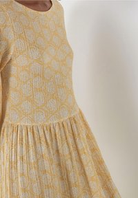 Vestido amarillo con un patrón texturizado de óvalos azules claro, escote redondo, mangas largas y una sección de falda fruncida.