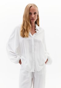 Weißes, knöpfbares Hemd mit Rüschkragen, aus leichtem Stoff gefertigt. Hat lockere Ärmel und zwei seitliche Taschen.