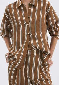 Camisa de botones con rayas verticales marrones y blancas, pantalones a juego de cintura alta, mangas remangadas, manos en los bolsillos, anillos plateados en los dedos.