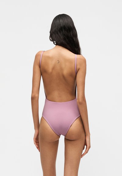 Femme aux longs cheveux foncés portant un maillot de bain une pièce mauve dos nu, debout de dos sur un fond clair uni.