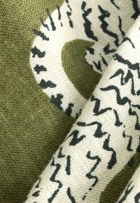 Textil, amely olajzöld és krémszínű színpalettával rendelkezik, texturált anyagból készült, és absztrakt fekete zigzag mintákkal díszített.