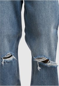 Světle modré džíny z denimu s potrhanými dírami na kolenou. Roztrhané okraje kolem děr a volný střih, který zdůrazňuje neformální styl.