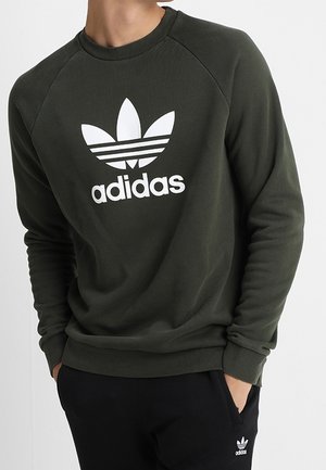 Person wearing dark green Adidas sweatshirt with white logo and black pants with small white Adidas logo, hands in pockets.