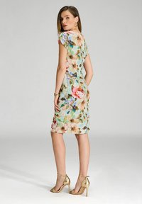 Robe florale avec de courtes manches capes, design ajusté en vert clair, ornée de grandes fleurs roses et bleues, associée à des sandales à talons hauts dorées.