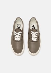 Une paire de baskets Vans en cuir taupe avec des lacets et des semelles blanches, vue de dessus sur un fond gris clair.