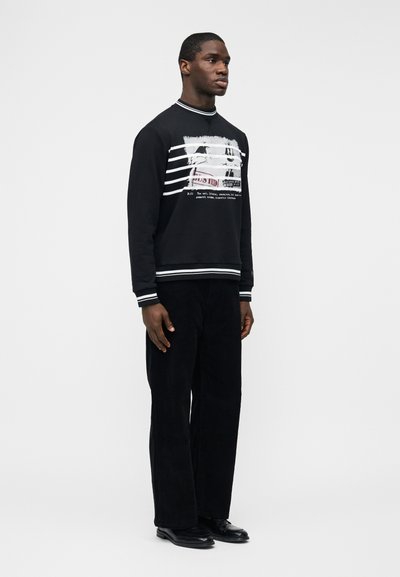 Homme debout, tourné vers la gauche, portant un sweat-shirt noir avec des rayures blanches et un motif graphique, un pantalon noir et des chaussures noires sur un fond blanc.