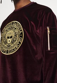 Camicia a maniche lunghe in velluto bordeaux con emblema di leone dorato e testo "GLORIOUS GANGSTA", dotata di tasca con zip sulla manica.