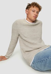 Beige gestrickter Pullover mit weitem Ausschnitt und langen Ärmeln, aus strukturiertem Stoff, über einem weißen Hemd getragen, kombiniert mit blauen Jeans.
