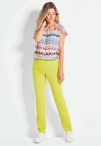 Mehrfarbiges Kurzarmoberteil mit abstraktem Muster, kombiniert mit leuchtend gelben geraden Hosen. Weiße Sneaker runden das Outfit ab.