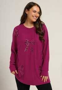 Sweater oversized magenta avec des motifs de nœud en sequins brillants, texture douce, manches longues et poches latérales, associé à un pantalon noir.