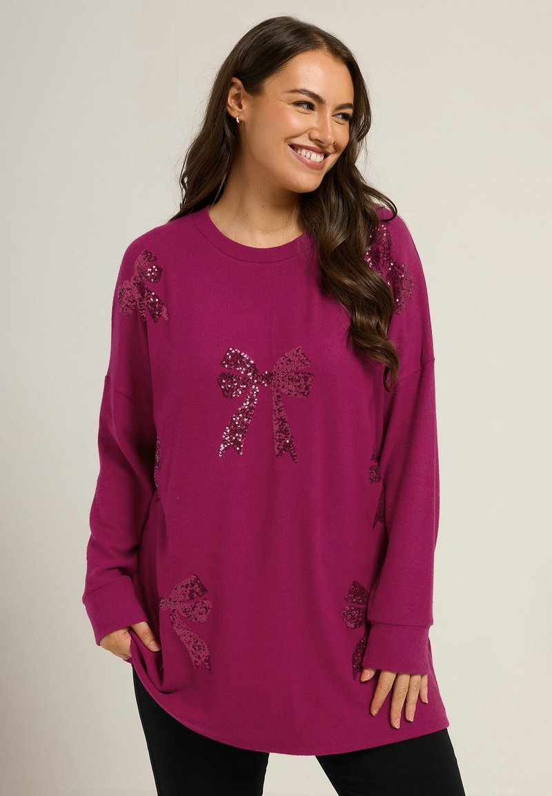 Sweater oversized magenta avec des motifs de nœud en sequins brillants, texture douce, manches longues et poches latérales, associé à un pantalon noir.