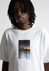 Biela bavlnená tričko s obdĺžnikovým grafickým potlačou motorkára na pozadí tmavého západu slnka. Krátke rukávy, okrúhly výstrih.