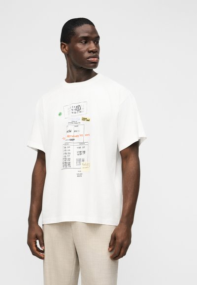 Άνδρας που φοράει λευκό φαρδύ T-shirt με εκτυπωμένο σχέδιο κάρτας βιβλιοθήκης και ανοιχτόχρωμο μπεζ παντελόνι, στέκεται μπροστά σε απλό φόντο.