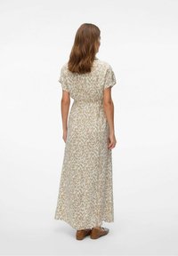Maxime dress in beige with a light pattern, short cap sleeves, elastic waist, and flowy fabric. Worn with brown slip-on shoes.