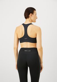 Sujeción deportiva negra con diseño de espalda nadadora, que presenta un pequeño logotipo rosa de Nike. Combinada con leggings negros de cintura alta que muestran un logotipo.