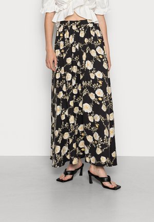 Femme portant une longue jupe noire à imprimé floral blanc et jaune, associée à des sandales à talons noires et un top court blanc.