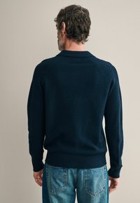 Sötétkék kötött pulóver bordázott aljjal és mandzsettával, gallérral és raglán ujjal. Világos mosású, textúrázott farmernadrággal párosítva.