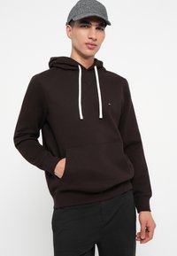 Mladý muž v tmavohnedej mikine s bielymi šnúrkami, s rukami v klokanke, so šedou vzorovanou baseballovou čiapkou a čiernymi nohavicami.