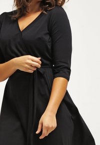Mustat kietaisumekko, jossa on kolmanneksen pituiset hihat, V-mallinen kaula-aukko ja solmittava vyö kyseisen vyötärölle, valmistettu sileästä kankaasta ja liukuvasta siluetista.