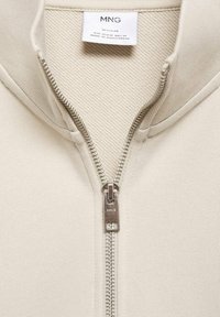 Prenda beige con cremallera y etiqueta de marca MNG que muestra el tamaño Regular, fabricada en Bangladés.