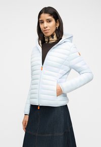 Γυναίκα που φοράει ελαφρύ μπλε καπιτονέ μπουφάν με κουκούλα, ριγέ ζιβάγκο και σκούρα τζιν φούστα, στέκεται με το ένα χέρι στην τσέπη του μπουφάν.