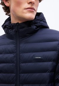 Νεαρό άτομο που φοράει σκούρο μπλε καπιτονέ μπουφάν με κουκούλα και το λογότυπο Calvin Klein στο στήθος, ορατό από το πηγούνι μέχρι το πάνω μέρος του κορμού.