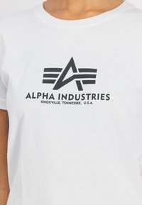 Balta medvilninė marškinė su juodu grafiniu logotipu, kuriame užrašyta "ALPHA INDUSTRIES" ir "KNOXVILLE, TENNESSEE, JAV." žemiau.