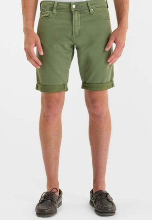 Uomo con pantaloncini verdi oliva arrotolati e scarpe da barca marrone scuro, in piedi contro uno sfondo chiaro e semplice.