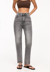 Femme portant un jean gris délavé taille haute, un crop top marron et des sandales plates noires à lanières, debout devant un fond blanc.